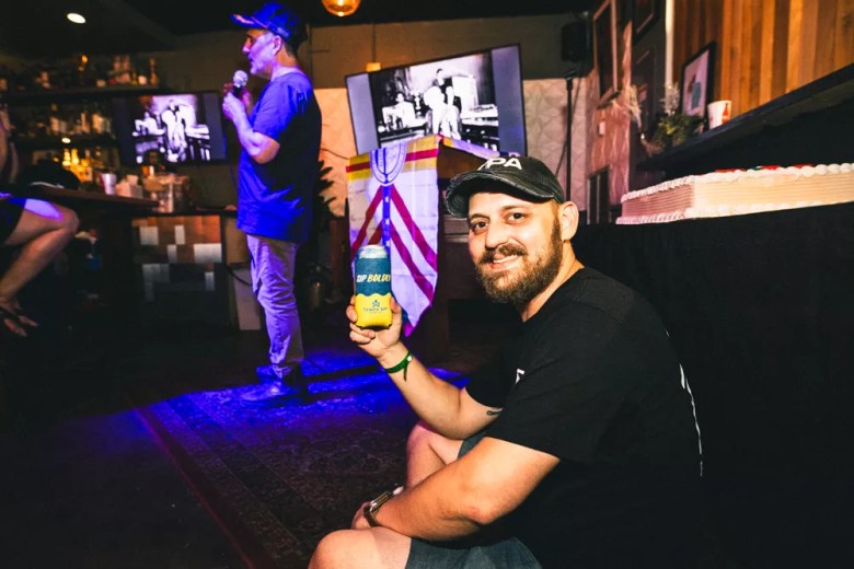 A man smiles holding up a beer next to someone speaking onstage at Tampa's Drunk History 2025