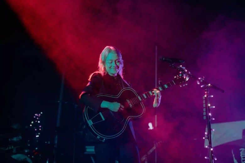 A concert photograph showing Phoebe Bridgers performing on stage during a concert at the Cuban Club in Ybor City, Florida, on May 24, 2022. Captured in a full-length shot, Bridgers stands centered on stage under dramatic red and purple spotlighting, smiling while playing an acoustic-electric guitar strapped across her shoulders. Her platinum blonde hair glows under the overhead lights against a hazy stage background decorated with fairy lights wrapping around microphone stands.
