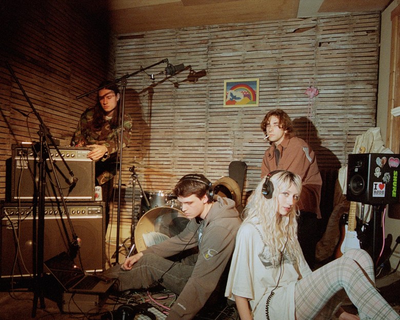 A promotional band photograph of the music group feeble little horse inside an unfinished room with exposed horizontal wooden lath walls. The four band members are casually positioned around various pieces of musical equipment. On the left, an individual with long dark hair, wearing a camouflage jacket, stands adjusting the knobs on a stack of Roland amplifiers. Towards the top right, a member with shaggy brown hair and a brown overshirt stands with a lit cigarette in their mouth. Seated on the floor in the center-left, a third member in a grey hoodie, grey pants, and black over-ear headphones looks down toward a laptop and a tangle of cables. On the bottom right, a fourth member with long blonde hair sits on the floor wearing a light-colored graphic t-shirt, plaid leggings, and over-ear headphones, looking directly at the camera. A drum kit is partially visible in the background, a small framed rainbow picture hangs on the wall, and a black speaker covered in audio brand stickers sits on the far right. The scene is lit with a direct flash, casting hard shadows on the textured wall behind them.