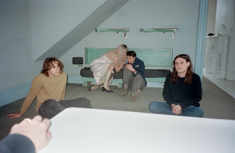 An indoor promotional photograph of the band feeble little horse inside a sparse room with pale blue walls, grey carpeting, and a slanted ceiling on the left. The image is shot with a direct flash, featuring a blurry hand and a large white rectangular surface partially obscuring the immediate foreground. The four band members are positioned casually around the space. On the far left, an individual with shoulder-length wavy brown hair wears a tan long-sleeve shirt and dark jeans, sitting on the floor and leaning back on one hand. In the center, a member with long blonde hair, wearing a light pink slip dress and tall laced boots, sits on a low, pale green bench and leans down to whisper into the ear of a third member. This third individual, wearing a dark blue hoodie, grey pants, and brown shoes, sits on the floor leaning against the bench to listen. On the right, a fourth member with long dark hair sits cross-legged on the floor wearing a black long-sleeve shirt and blue jeans, looking directly at the camera.