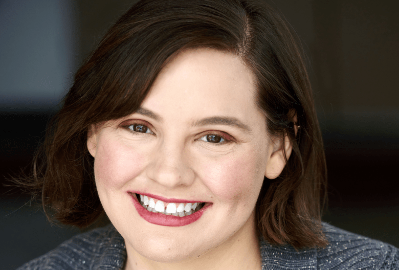 A headshot photograph of improv comedian Sarah Schultz smiling warmly at the camera. Positioned center frame in a medium close-up shot, Schultz features brown shoulder-length hair styled with a side part. They wear a deep purple v-neck top layered under a textured dark grey open knit cardigan sweater, set against a softly blurred, dark neutral background.
