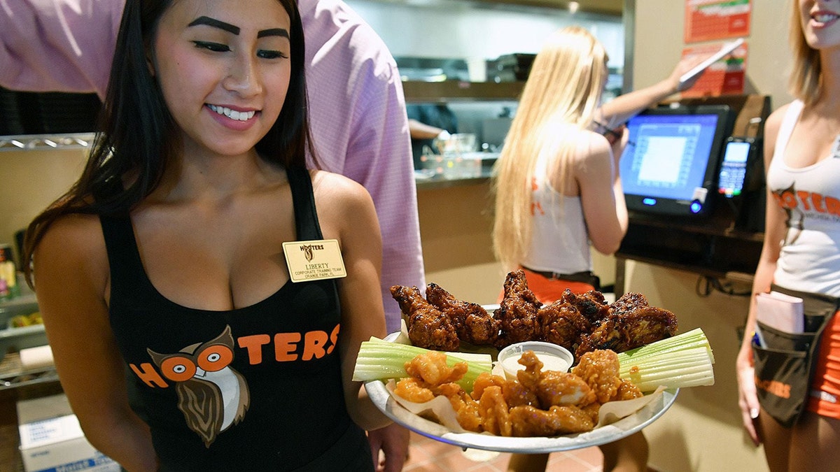 A Hooters waitress wearing a black top holds a plate of chicken wings. In the background are two Hooters waitresses in the traditional white and orange uniforms.