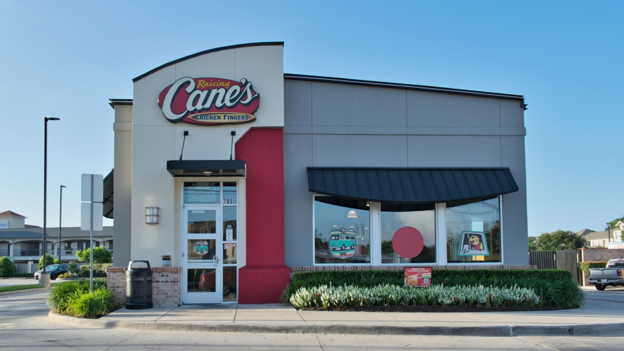 Exterior of a Raising Cane's Chicken Fingers restaurant with the chain's signature red-and-white branding, large front windows and landscaped entrance.