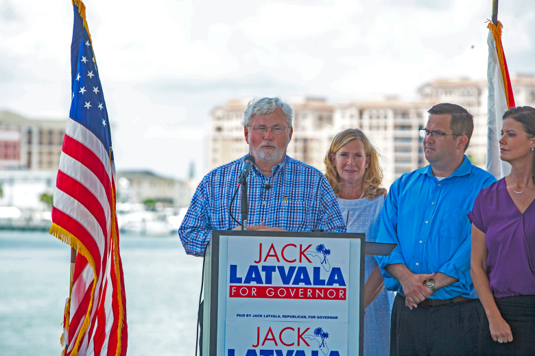 Florida Representative Chris Latvala wearing a blue button-down shirt and glasses, standing on an outdoor stage next to a woman in a purple shirt during a political campaign event in Clearwater.