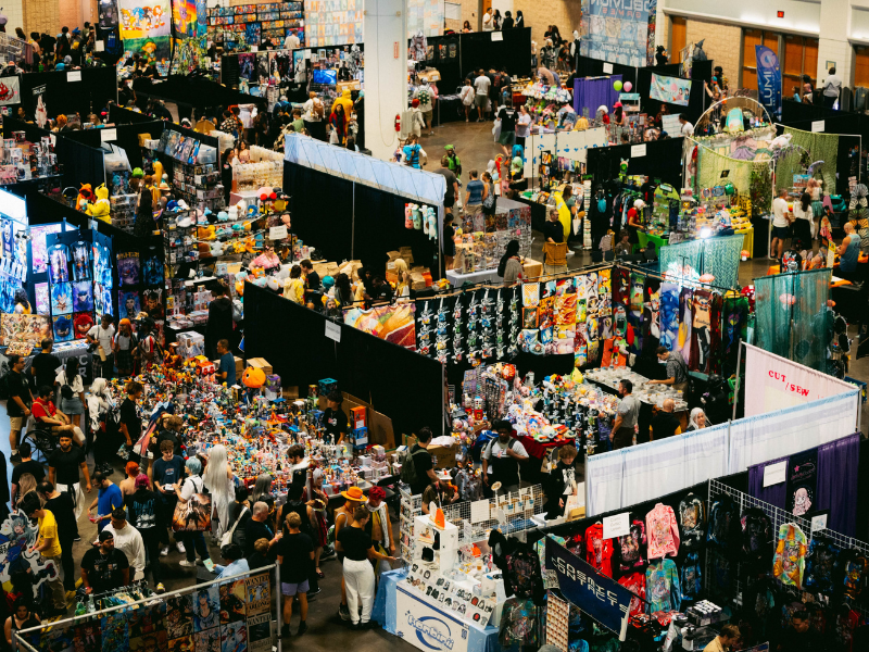 Exhibit hall during METROCON 2025 at Tampa Convention Center filled with vendor booths selling merch such as toys and clothing as attendees in costumes walk around.