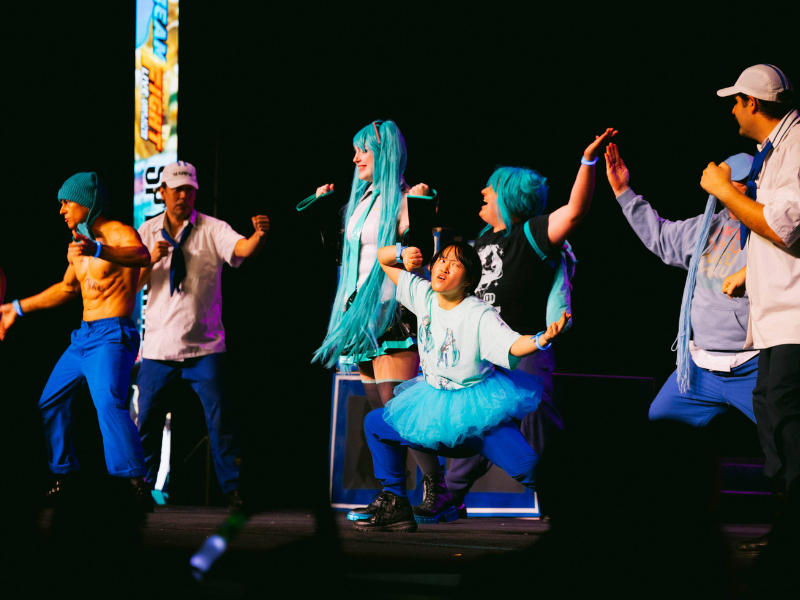 Group of people in costumes including blue hair and a blue tutu stand on stage in various poses during METROCON 2025.