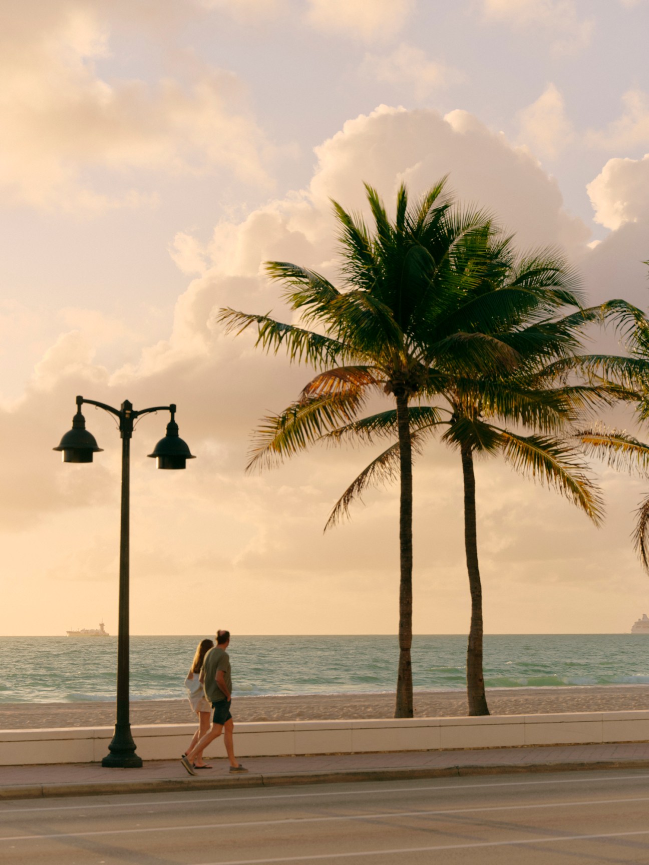 Couple walking along North Fort Lauderdale Beach Boulevard waterfront promenade with palm trees and ocean view at sunset