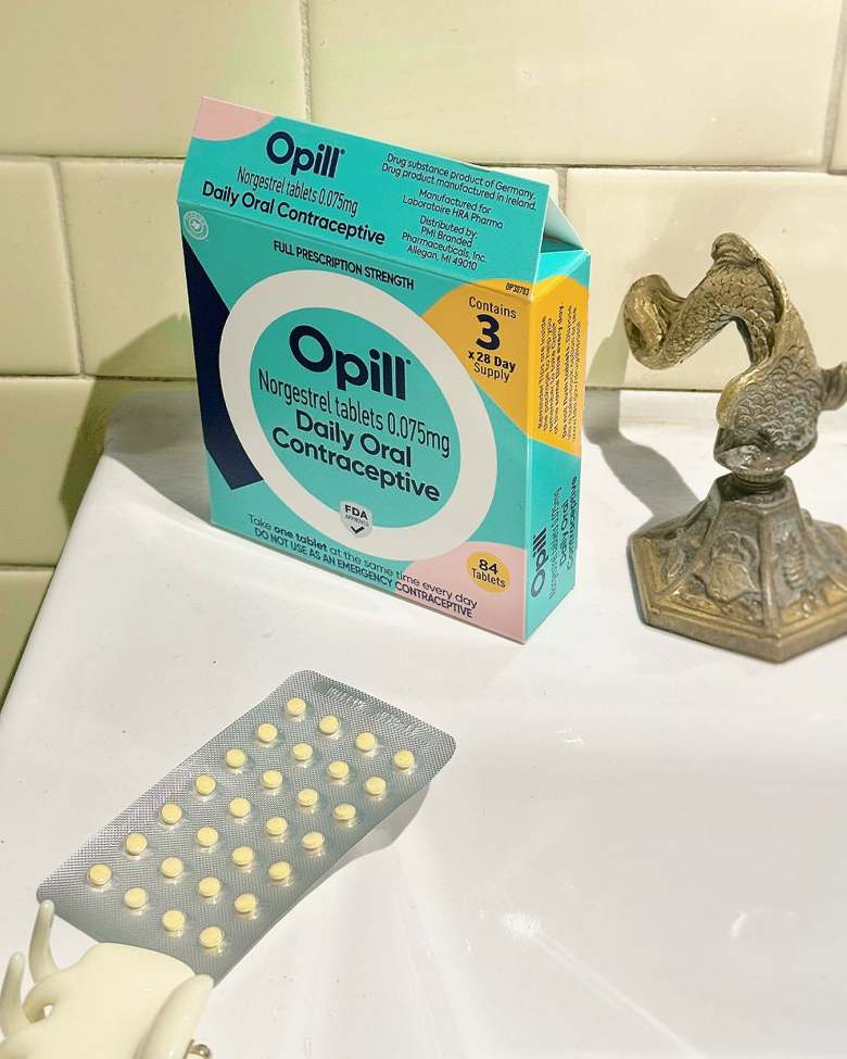A photograph of Opill over-the-counter birth control pills sitting on a white porcelain bathroom sink against a beige tiled wall background. In the foreground, a grey textured blister pack contains a grid of 28 small, round, yellow pills. Behind it stands the vibrant turquoise, orange, and pink cardboard packaging box, clearly displaying the text "Opill Norgestrel tablets 0.075mg Daily Oral Contraceptive" and noting a "3-month supply." To the right of the box sits a small, decorative metallic sculpture of a leaping fish on a hexagonal base.