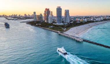 Aerial view of a yacht sailing near a beach with high-rise buildings, a sandy shoreline, and the ocean at sunset in a coastal city—an iconic scene that captures the vibrant allure of South Florida Real Estate.