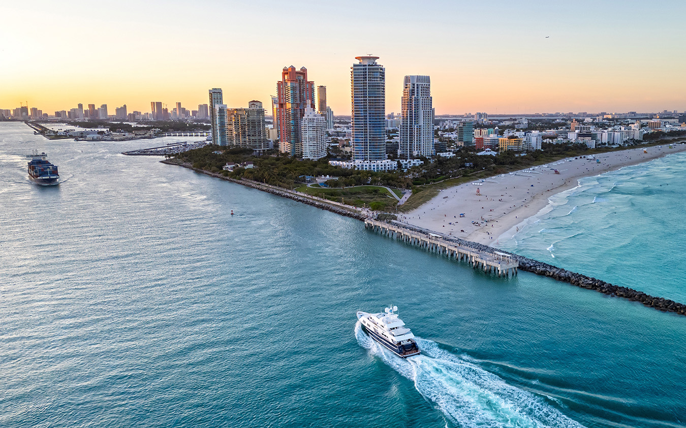 Aerial view of a yacht sailing near a beach with high-rise buildings, a sandy shoreline, and the ocean at sunset in a coastal city—an iconic scene that captures the vibrant allure of South Florida Real Estate.