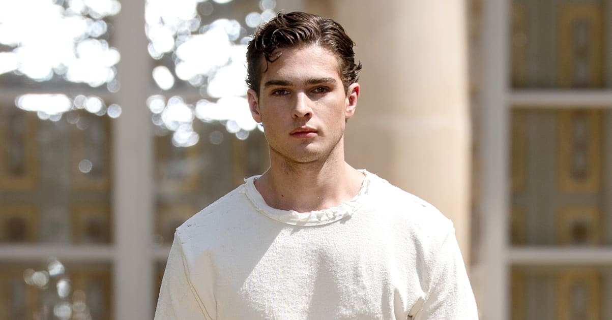 Clavicular in a white outfit walking outdoors, with a neutral expression and styled hair.