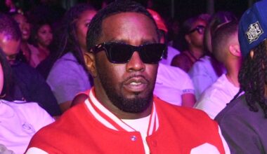 Diddy in sunglasses and a red varsity jacket sits among a crowd at an event.