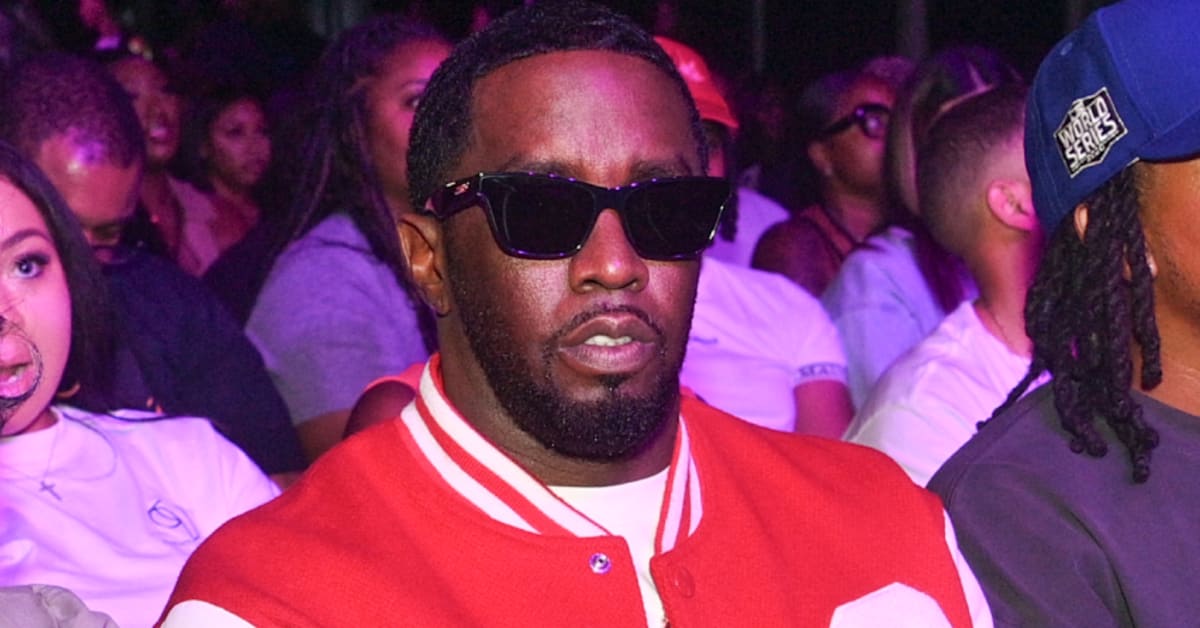 Diddy in sunglasses and a red varsity jacket sits among a crowd at an event.