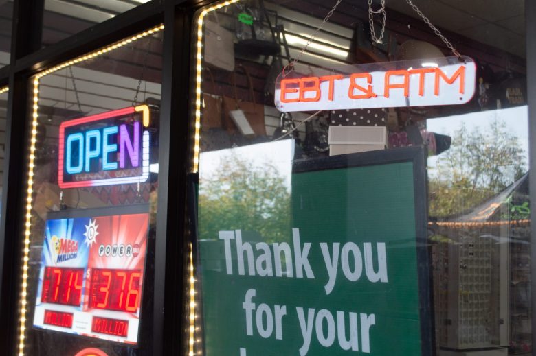 Neon 'OPEN' and 'EBT & ATM' signs glowing in a convenience store window next to lottery jackpot displays and a thank you sign.