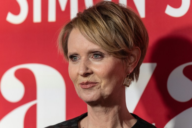 Cynthia Nixon posing against a red background with white letters