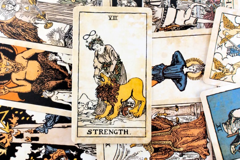 A flat-lay photograph showcasing a spread of classic Rider-Waite tarot cards scattered on a flat surface, with the "Strength" Major Arcana card featured prominently in vertical orientation at the center. The Strength card, labeled with Roman numeral "VIII" at the top and "STRENGTH" at the bottom, depicts a figure in a white robe crowned with a flower wreath and an infinity symbol floating above their head, calmly taming a yellow lion under a yellow sky. Other major and minor arcana cards—including The Devil and The Hanged Man—lie partially overlaid and angled beneath the central card in a collage arrangement.
