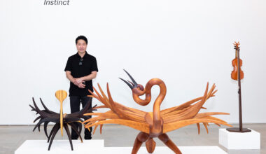 A man stands behind three works of art made from wood, including two decorative chairs and a violin.