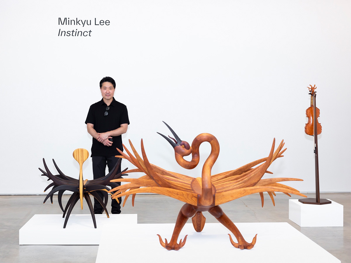 A man stands behind three works of art made from wood, including two decorative chairs and a violin.