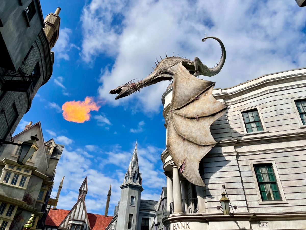A dragon sculpture on a building facade breathes fire against a backdrop of blue sky and other fantasy-themed buildings, reminiscent of the magical world you can explore with a Universal Orlando Resort ticket.