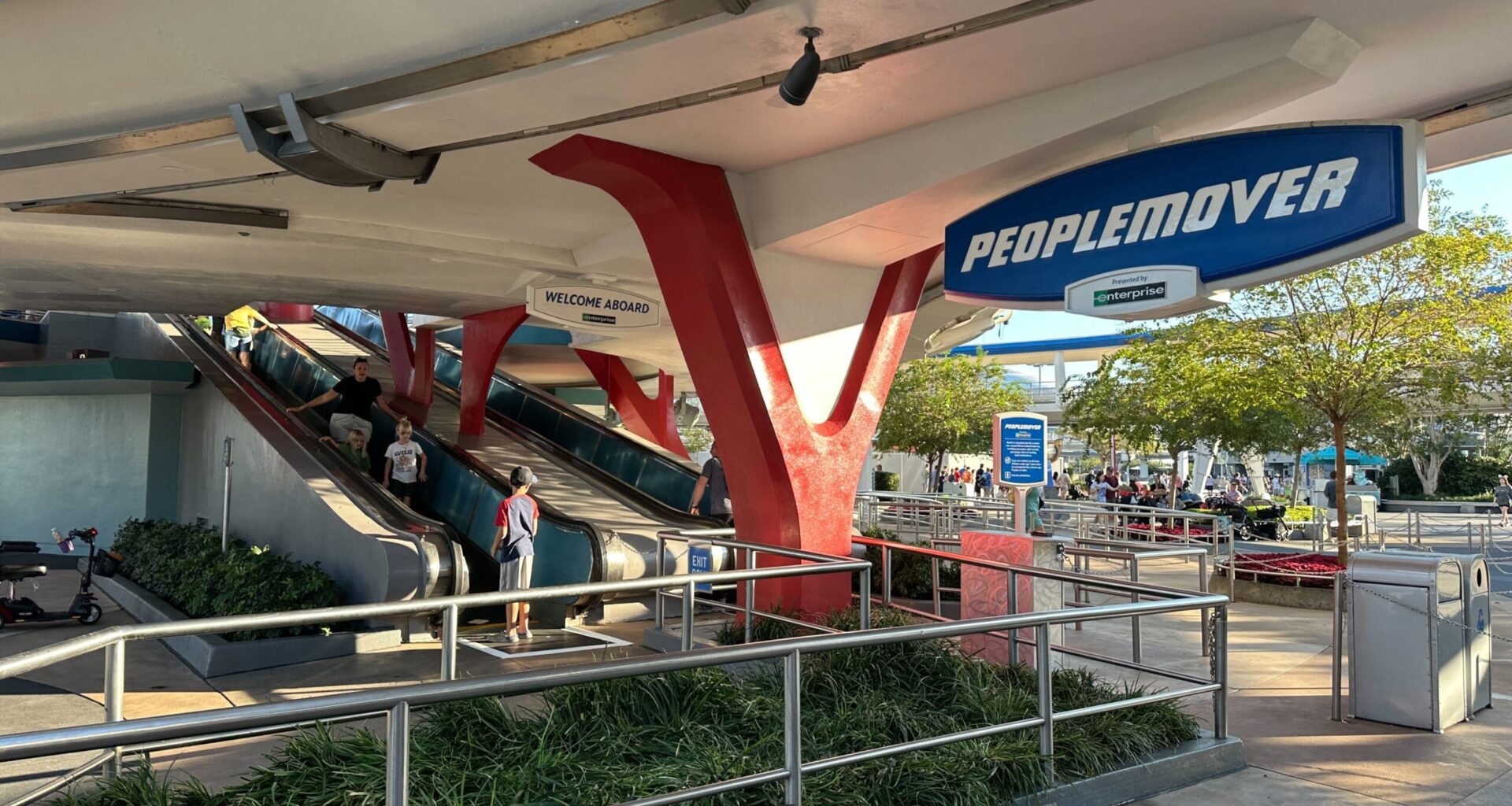 Two escalators lead to the PeopleMover ride entrance under a white canopy with blue and red accents at a theme park.