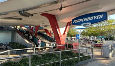 Two escalators lead to the PeopleMover ride entrance under a white canopy with blue and red accents at a theme park.