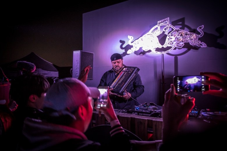 A photograph by Sonstar showing electro hip-hop musician Egyptian Lover performing live on stage at Some Sevit during the Red Bull Music Academy Bass Camp in Seoul, South Korea, on September 29, 2017. Captured in a medium shot under dramatic purple stage lighting, Egyptian Lover leans forward over a DJ setup holding a Roland synth controller. Behind him, a illuminated metallic Red Bull logo sculpture mounts against the wall. In the shadowed foreground, concertgoers raise their smartphones to record the performance in the dim venue interior.