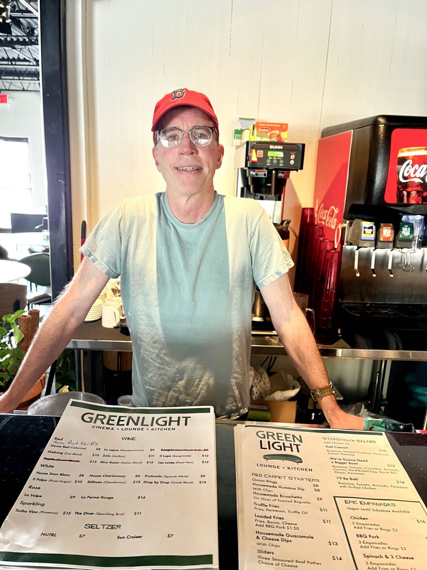 Man with a blue baseball cap and glasses, wearing a green t-shirt and standing in front of menus