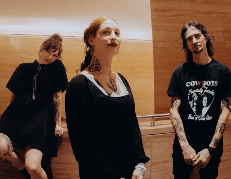 A promotional band photograph by Mack Knox showing Kirsten Clauser, Nikki Raven, and Alton Plemmons of Hovercar posing inside a modern room with warm wood-paneling. Captured in a medium wide shot, the three band members stand side-by-side from left to right: Kirsten Clauser leans against a wooden ledge wearing a black short-sleeved collared dress and bolo tie with dark tattoos visible on her arms and leg; Nikki Raven stands center frame wearing a long-sleeve black top with a square white neckline, dark red lipstick, and a neck tattoo; and Alton Plemmons stands on the right wearing a black graphic t-shirt reading "COWBOYS Are Frequently Secretly Fond of Each Other" with sleeve tattoos on both arms under warm, indirect lighting.