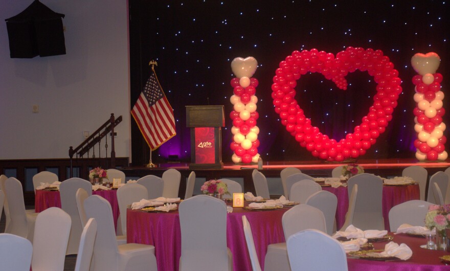 A ballroom is laid out with white chairs and pink-topped tables that a face a stage occupied by pink-and-white, heart-shaped balloons and a podium with a "GTE Financial" overlay. An American flag stands at the edge.