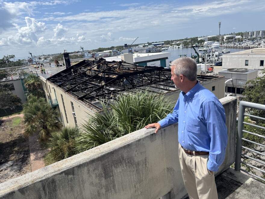 Man wearing khaki slacks and a blue long sleeve collared shirt with his hand on concrete balcony of an outdoor stairwell with his face looking toward a burned building which has some surrounding green trees. Beyond that is a blue sky with white clouds, boats docked in a blue water harbor.