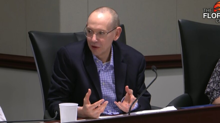A bald man in checked blue shirt and blazer gestures before a mic