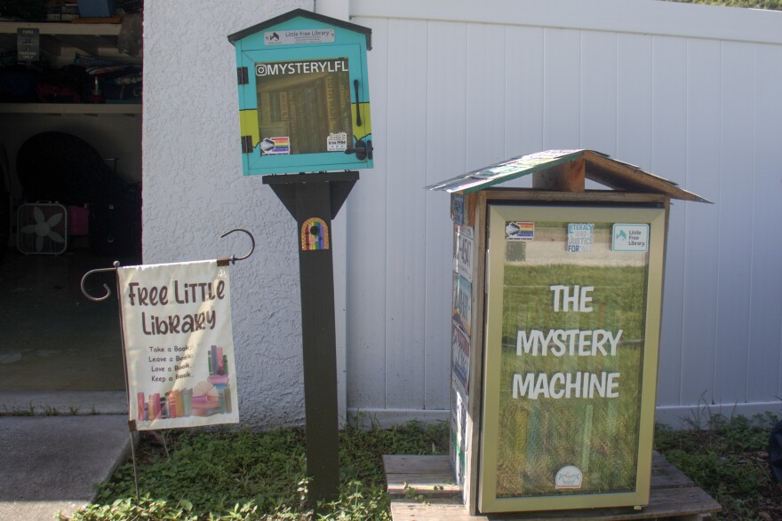 A little case full of books standing on a wooden pole in front of a white fence. One one side is a little white flag that reads "Free Library," and on the other is a minifridge full of books, covered in licenses plates.