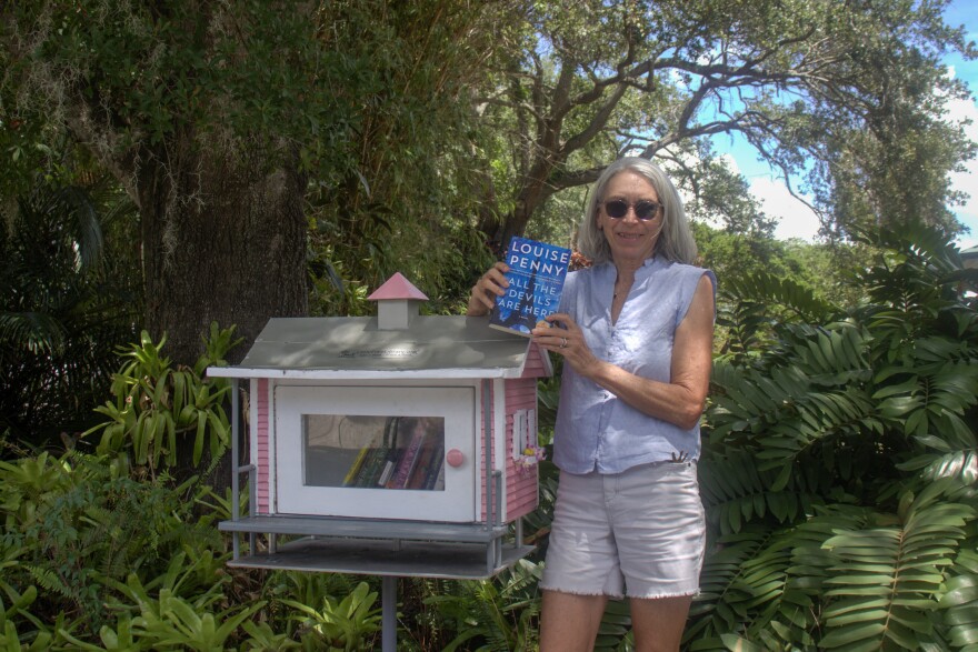 A woman wearing sunglasses and holding up a blue book stands next to a little case of books that resembles a small pink house.