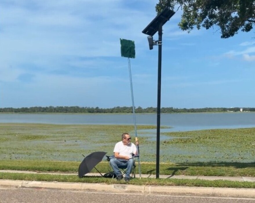 Older man sits by road blocking a Flock camera with a large pole and sign on it 