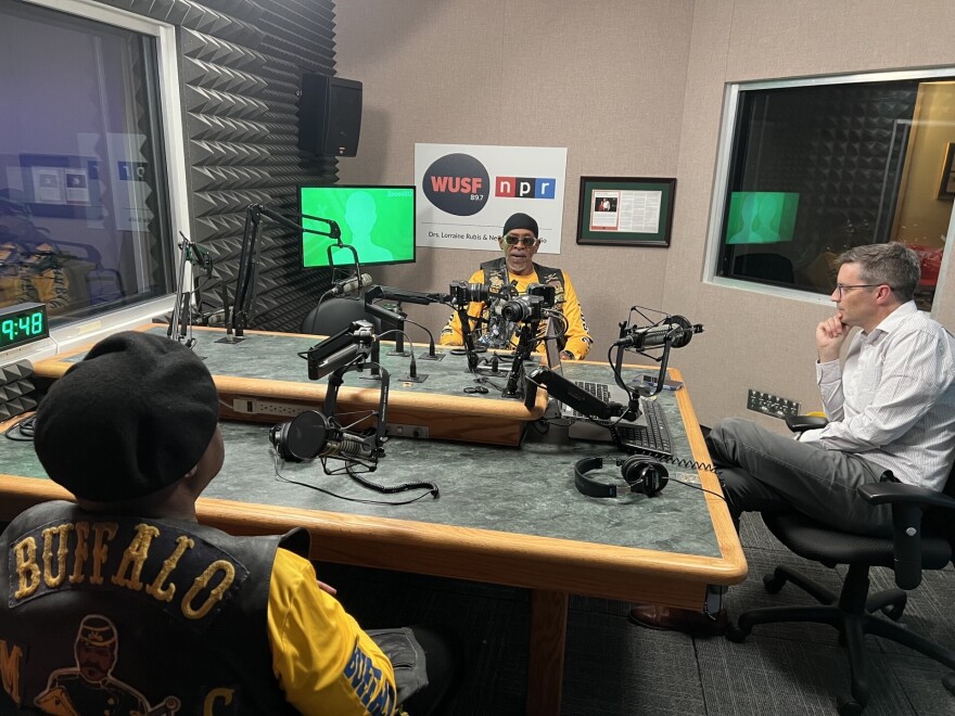 Two Black men in yellow shirts and leather vests sit at mics in a studio with the host, a white man.
