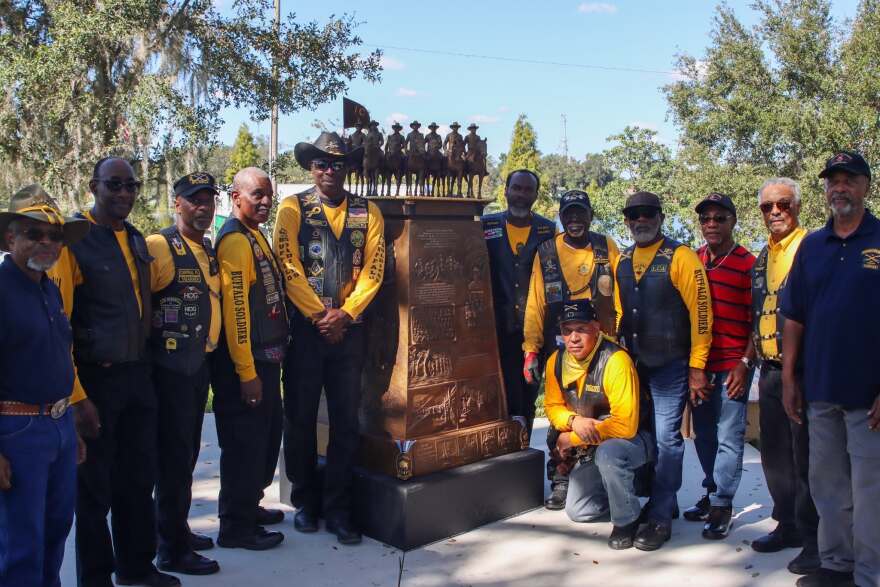 A group of Black men and women crowd around a bronze statue. They’re wearing mustard yellow long sleeved shirts and black leather vests adorned with patches and pins.