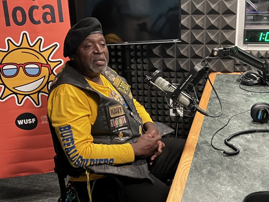 A Black man in a yellow shirt and leather vest sitting at a mic in a studio.