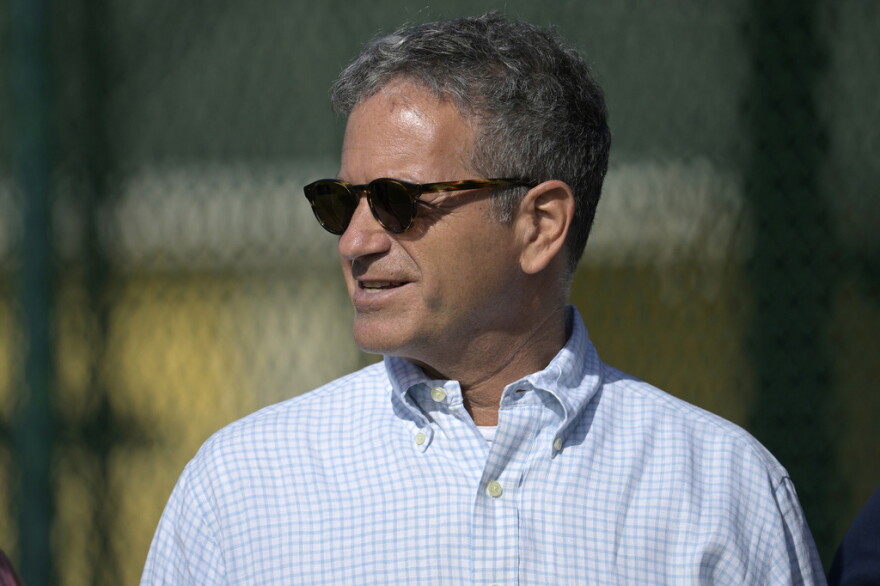 Man with salt-and-pepper hair with sunglasses, wearing a blue and white dress shirt and smiling, looking to the left