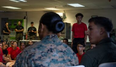 A military briefing or orientation session is taking place indoors, with uniformed Marines addressing a group of young civilians wearing red shirts. The scene appears to be a youth outreach or recruitment event, possibly a Junior ROTC or similar program. A Marine in camouflage uniform is speaking to seated participants while others stand and observe.