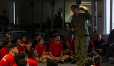 A military personnel member in olive/flight suit uniform addresses a group of young people seated on the floor, all wearing matching red t-shirts with what appears to be Marines branding. The setting appears to be an indoor briefing or orientation session, likely a recruiting or youth outreach event. Additional military personnel are visible in the background observing the presentation.