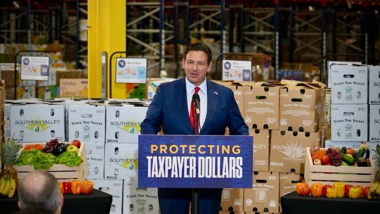 Florida Governor Ron DeSantis speaking at a podium with a "Protecting Taxpayer Dollars" sign in a warehouse surrounded by produce boxes and fresh vegetables.