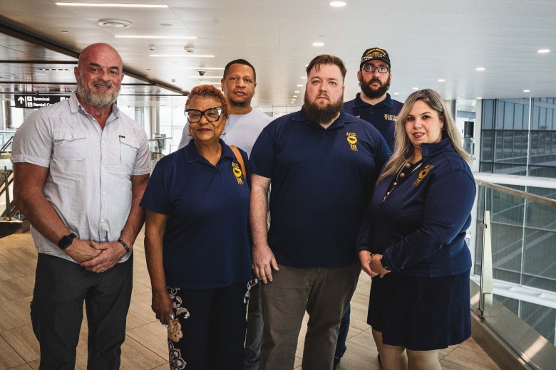 A indoor group photograph by Dave Decker showing six members of the American Federation of Government Employees at Tampa International Airport on August 6, 2026. The group stands together on an interior walkway, consisting of four men and two women. In the front row from left to right, a man in a light grey short-sleeved button-down shirt and dark trousers stands beside a woman in a navy blue short-sleeved polo shirt with an AFGE emblem and glasses, a man in a navy blue polo shirt with an AFGE logo, and a woman in a long-sleeved navy blue collared jacket and dark skirt. Two men stand behind them in the back row, one in a light grey shirt and another wearing a dark cap and navy polo shirt, set against large glass windows overlooking an airport terminal concourse.