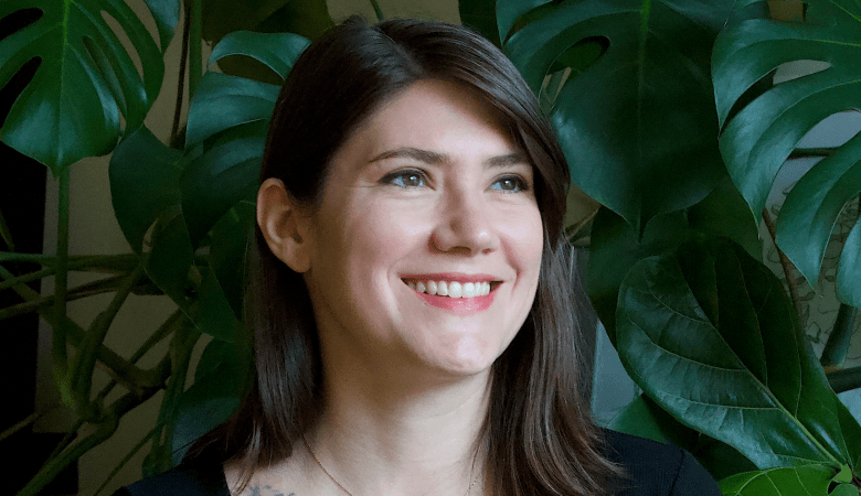 Alt-text: A close-up photograph by Scott Krier showing Arielle Hebert smiling warmly in front of lush indoor foliage. Captured in a medium close-up headshot, Hebert looks toward the right of the frame with a wide, bright smile, featuring shoulder-length dark brown hair parted on the side, a delicate necklace, and a black top. She is positioned against a vibrant background of large green monstera and fiddle-leaf fig leaves.