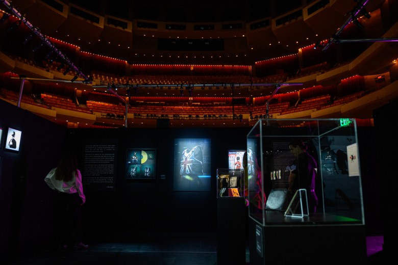 A wide interior photograph showing a preview of "The Art of Banksy" exhibition staged inside Morsani Hall at the David A. Straz Center for the Performing Arts in Tampa, Florida, on July 30, 2026. Set under low ambient gallery lighting, the stage of the dark, multi-tiered auditorium is transformed into a walk-through art gallery. In the foreground, glass display cases hold authenticated Banksy artifacts, flanked by illuminated wall-mounted artworks including a glowing central piece, framed prints, and informative black text panels along the gallery walls, while a visitor stands observing the installation.