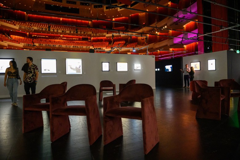 A wide-angle interior photograph showing a different angle of "The Art of Banksy" exhibition inside Morsani Hall at the David A. Straz Center for the Performing Arts in Tampa, Florida, on July 30, 2026. Set against the background of the auditorium's red-lit, multi-tiered seating balconies, the foreground features a dark polished floor arranged with plush velvet armchairs and dark tables. Along white temporary partition walls, backlit framed artworks and digital screens are displayed under purple and ambient overhead lighting, while visitors walk through the gallery space viewing the exhibits.