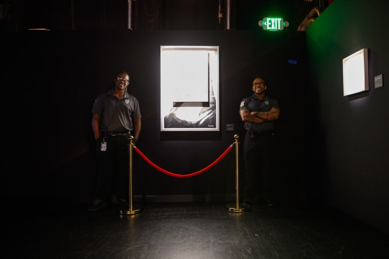 A photograph showing two security guards standing alongside a framed artwork at "The Art of Banksy" exhibition inside Morsani Hall at the David A. Straz Center for Performing Arts in Tampa, Florida, on July 30, 2026. Captured against a dark, shadowy gallery wall under green EXIT sign illumination and accent lighting, two guards in dark uniforms stand on either side of a bright, illuminated framed artwork framed by a velvet rope stanchion in the center. Another framed artwork hangs illuminated on the adjacent dark wall to the right.