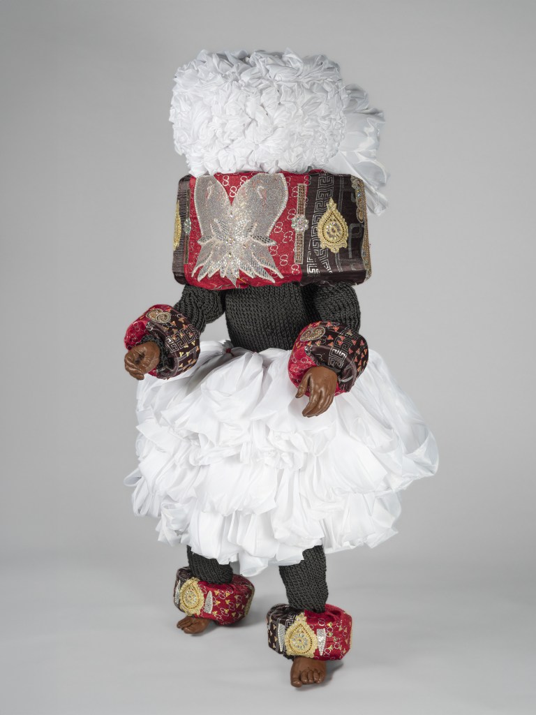 A studio photograph of the full life-size Afia Awan Masquerade Ensemble artwork created in 2022 by Chief Ekpenyong Bassey Nsa, held in the collection of the New Orleans Museum of Art. Displayed against a neutral light grey backdrop, the figure is dressed in a layered masquerade costume featuring a large, textured white headpiece, a wide collar trimmed in red fabric with intricate metallic gold embroidery, and a voluminous white ruffled skirt. The dark arms, legs, and bare feet are detailed with matching embroidered red-and-gold cuffs at the wrists and ankles.