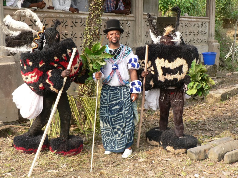 A photograph showing Sheku 'Goldenfinger' Fofanah standing between two masqueraders during the Massaboni Ordehlay procession in Lunsar, Sierra Leone, on December 26, 2022. Captured outdoors in a full-length wide shot, Fofanah stands centered wearing a black top hat, a patterned blue-and-white waist wrap, white sneakers, and arm bands while holding a wooden staff and green leafy branches. He is flanked on both sides by elaborate Mami Wata masquerade figures wearing dark, heavy raffia suits, tall horned headpieces with white feathers, and ankle cuffs while holding tall wooden staffs against an outdoor village backdrop.