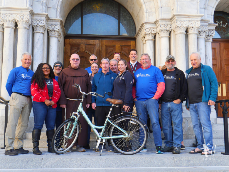 A group photograph showing members of Sacred Heart Catholic Church's "Bikes from the Heart" ministry gathered outside Sacred Heart Catholic Church in downtown Tampa, Florida, on February 21, 2025. Positioned on the exterior stone steps under arched marble doorways, a group of about twelve volunteers—wearing blue long-sleeved t-shirts, jackets, and casual jeans—pose together behind a light blue step-through bicycle parked on a kickstand in the center foreground. A Franciscan friar in a traditional brown habit stands near the center left among the volunteers under natural daylight.
