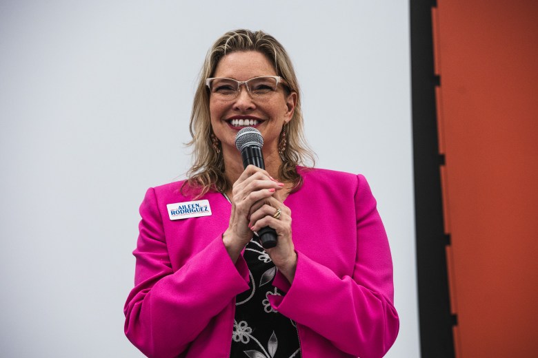 A photograph by Dave Decker showing Aileen Rodriguez speaking at the C. Blythe Andrews Jr. Public Library in Tampa, Florida, on July 9, 2026. Captured in a medium shot, Rodriguez stands smiling behind a microphone held with both hands, wearing a bright magenta pink blazer over a black-and-white floral print top, cat-eye eyeglasses, and a name tag reading "AILEEN RODRIGUEZ" pinned to her lapel against a light, plain backdrop.